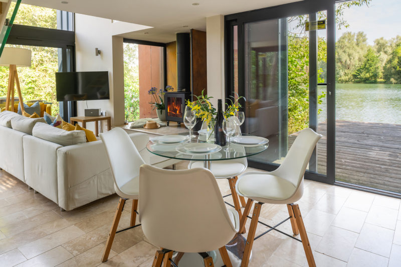 Modern holiday home living room and dining area with glass table, white chairs, and large windows leading to a wooden deck with a lake view in the Cotswolds.